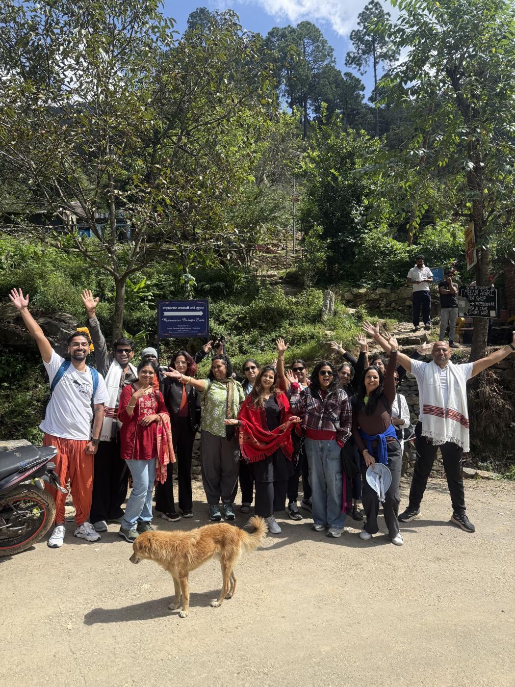 A WithShambhavi Yatra group smiling and waving together on a sunlit mountain path, with a dog in the foreground.
